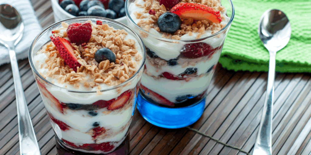 2 clear glasses layered with vanilla yogurt, granola, blueberries and strawberries set on a wooden table accompanied by a silver spoon